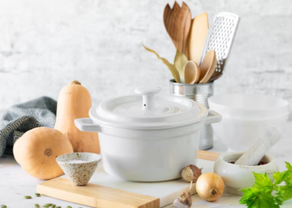 a white Dutch oven on a countertop, with onions, a mortar and pestle, and other kitchen utensils.