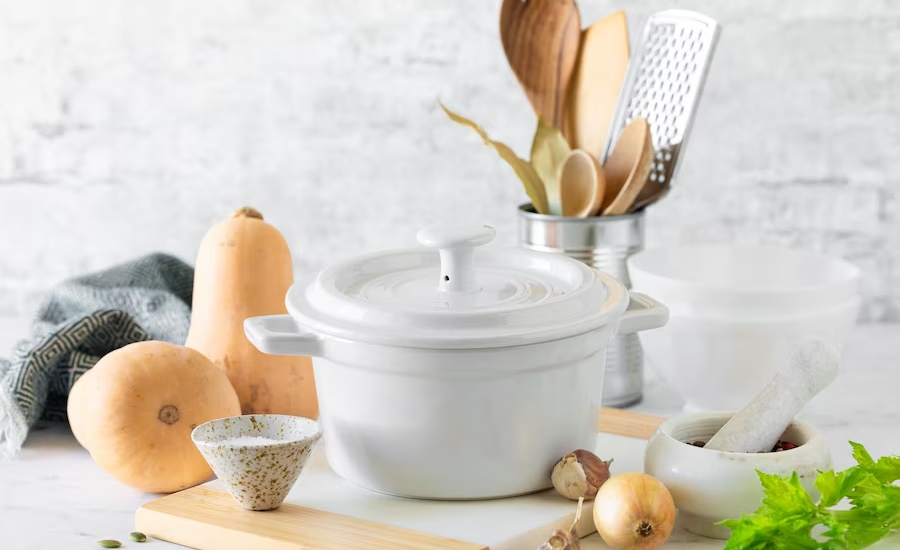 a white Dutch oven on a countertop, with onions, a mortar and pestle, and other kitchen utensils.